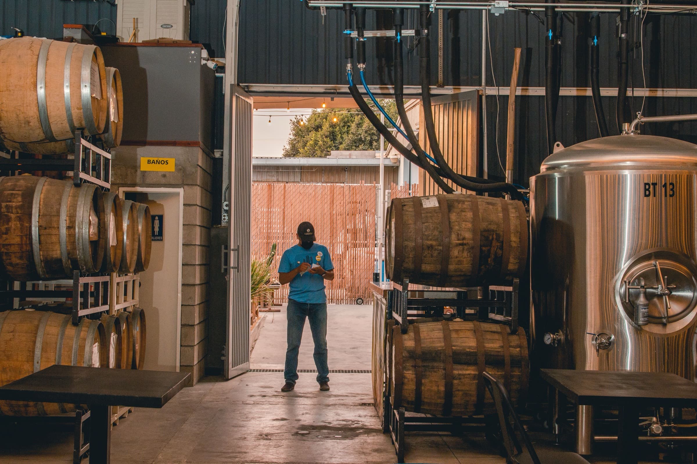 A brewery floor with stacked oak barrels, stainless fermentation tanks and a brewer checking stock