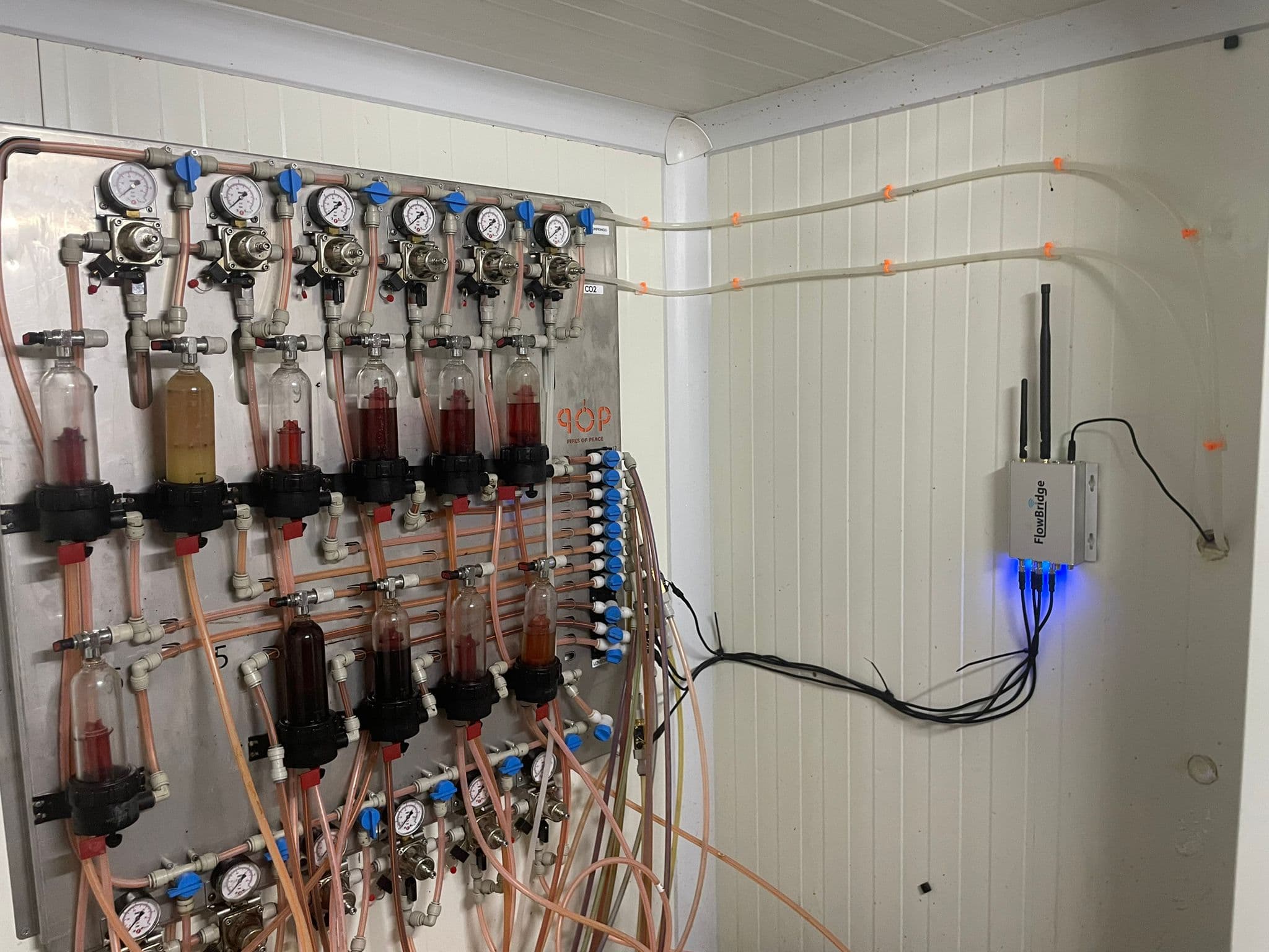 A FlowBridge gateway mounted on a cellar wall with its indicator lights lit and aerials up, beside a gas manifold board and beer lines