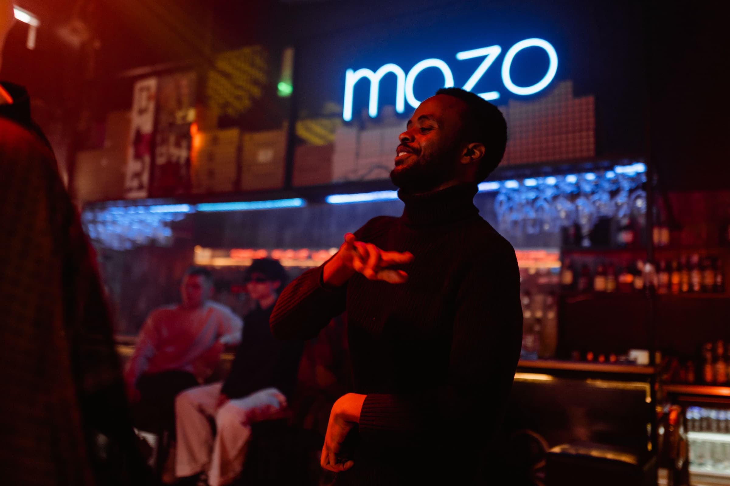 A bar at night, back-lit spirits and glassware, customers seated at the counter